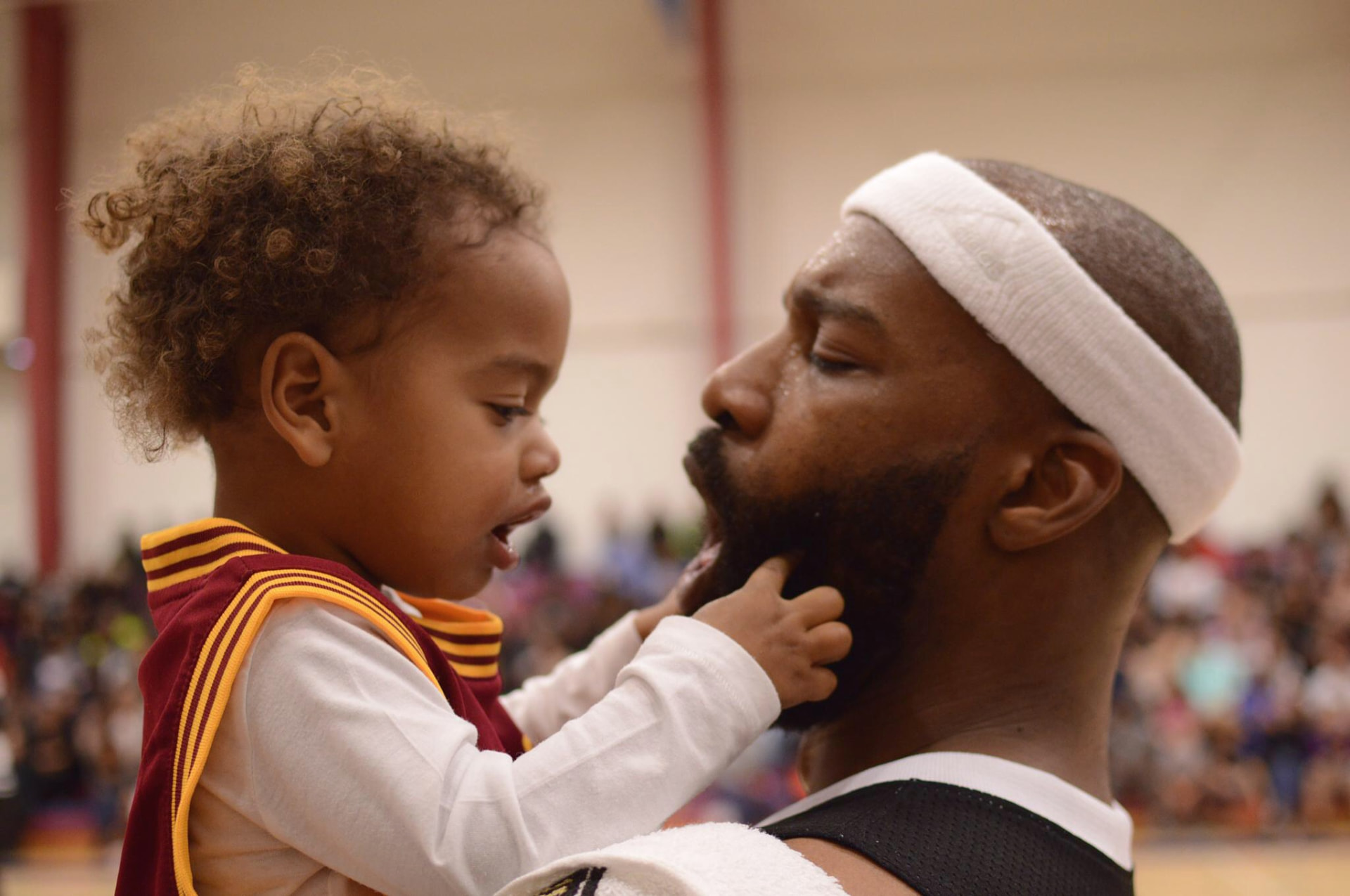 A basketball player in a white headband smiles as a young child reaches toward his beard at the event.