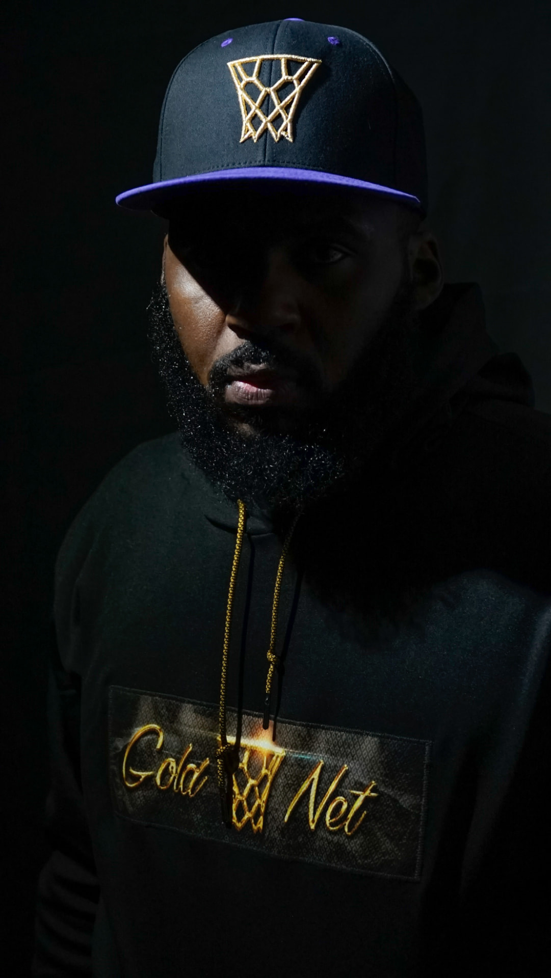 A low-key portrait of a man wearing a basketball hoop cap and Goldnet hoodie.