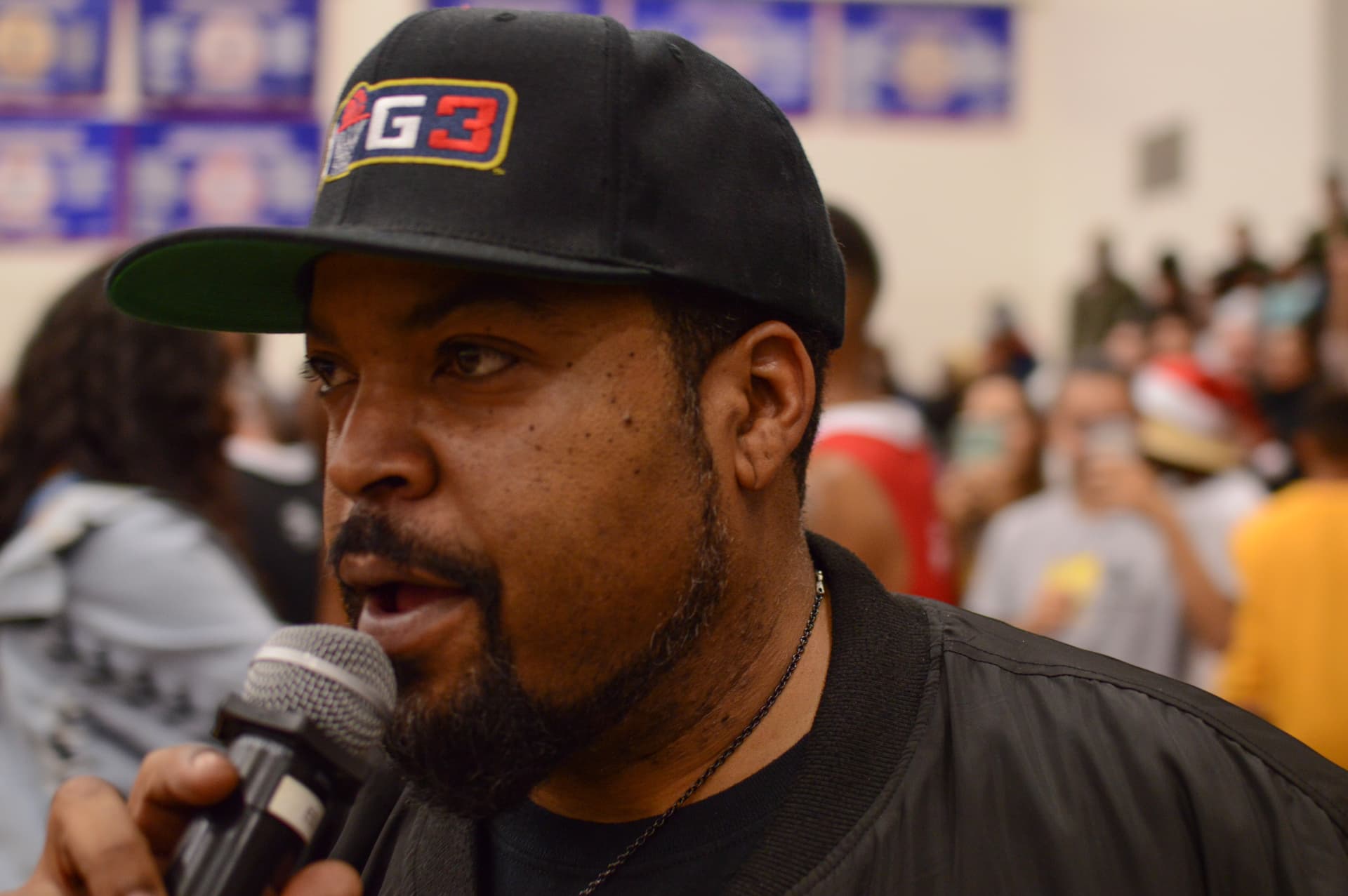 A speaker wearing a Big3 cap speaks into a microphone inside a crowded basketball gym.